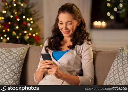 winter holidays, technology and people concept - smiling woman with smartphone at home over christmas tree lights on background. smiling woman with smartphone at home on christmas