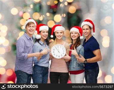 winter, holidays, time and people concept - group of smiling teenagers in santa helper hats with clock pointing finger over christmas tree lights background