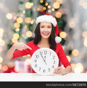 winter, holidays, time and people concept - smiling woman in santa helper hat and red dress with clock over christmas tree and ligths background