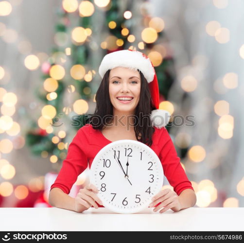 winter, holidays, time and people concept - smiling woman in santa helper hat and red dress with clock over christmas tree lights background