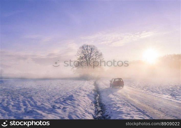 Winter image with the silhouette of a car driving on a snowy country road, through mist and the sunrise light.