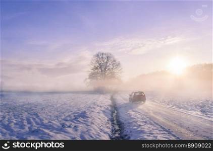 Winter image with the silhouette of a car driving on a snowy country road, through mist and the sunrise light.