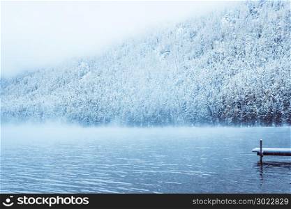 Winter lake with a pier for boats. Snow-covered pine trees on the shore. The Altai mountains, fog over the winter lake.