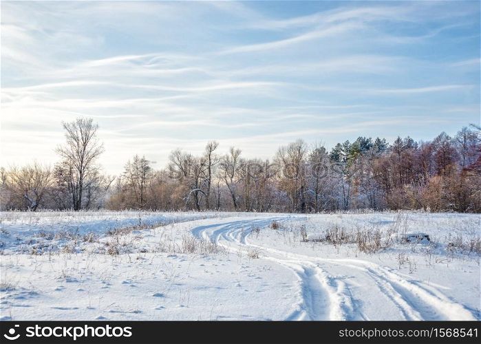 Winter landscape with a road on the white snowy expanse and a forest against the blue sky with light wavy clouds
