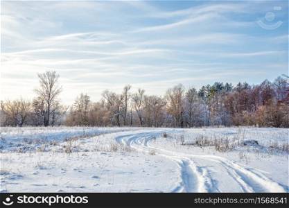 Winter landscape with a road on the white snowy expanse and a forest against the blue sky with light wavy clouds
