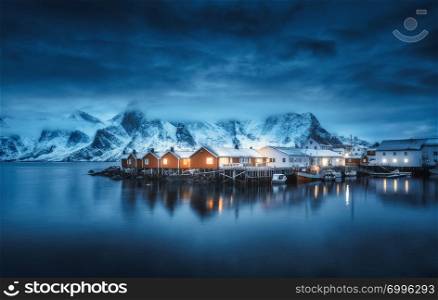Winter landscape with houses, illumination, snowy mountains, sea, blue cloudy sky reflected in water at night. Small fishing village with boats at twilight, Lofoten islands, Norway. Norwegian rorbu