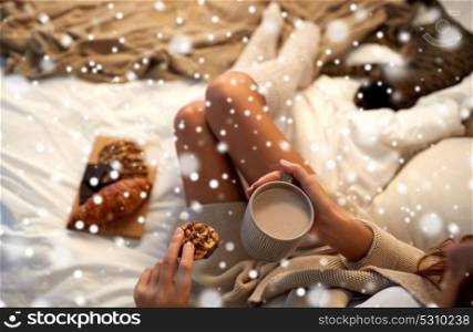 winter, leisure and people concept - close up of young woman with cup of coffee or cacao and cookie in bed at home over snow. close up of woman with cocoa cup and cookie in bed