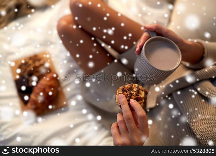 winter, leisure and people concept - close up of young woman with cup of coffee or cacao and cookie in bed at home over snow. close up of woman with cocoa cup and cookie in bed