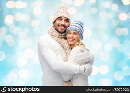 winter, love, couple, christmas and people concept - smiling man and woman in hats and scarf hugging over blue holidays lights background