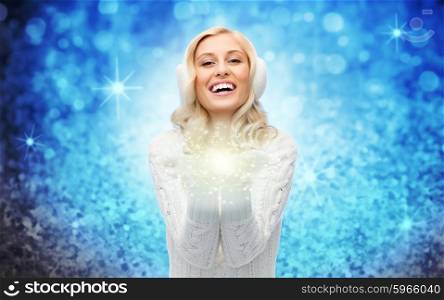 winter, magic, christmas and people concept - smiling young woman in earmuffs and sweater holding fairy dust on her palms over blue glitter or lights background