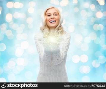 winter, magic, christmas and people concept - smiling young woman in earmuffs and sweater holding fairy dust on palms over blue holidays lights background