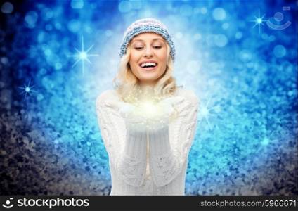 winter, magic, christmas and people concept - smiling young woman in hat and sweater holding fairy dust on her palms over blue glitter or lights background