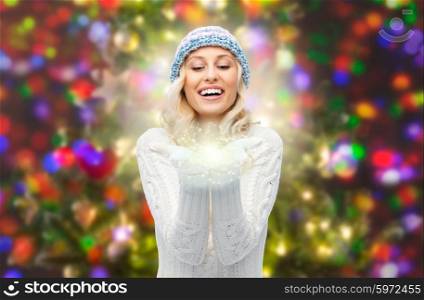 winter, magic, christmas and people concept - smiling young woman in hat and sweater holding fairy dust on her palms over lights background
