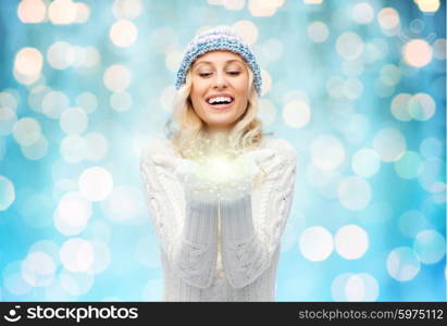 winter, magic, christmas and people concept - smiling young woman in winter hat and sweater holding fairy dust on palms over blue holidays lights background