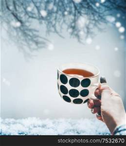 Winter mood concept. Female hand holding cup of hot tea at cold snow winter landscape nature background