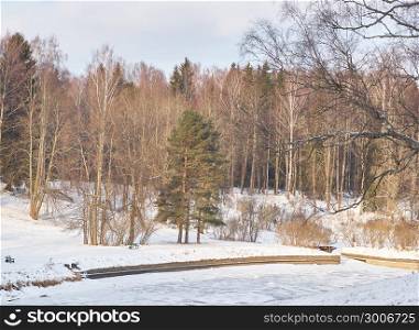 Winter park. River and trees and a bench on the beach under the snow