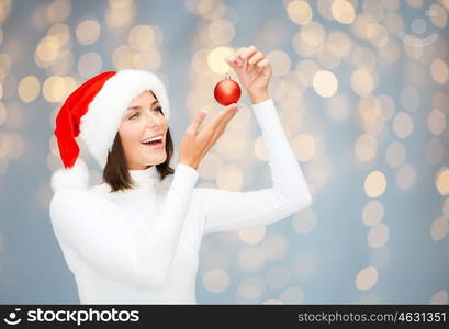 winter, people and holidays concept - happy smiling woman in santa helper hat with christmas tree decoration ball over lights background