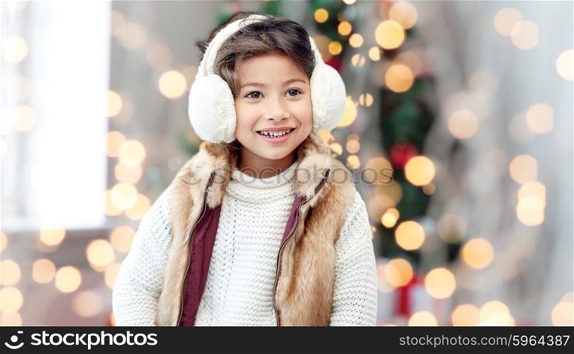 winter, people, childhood and happiness concept - happy little girl wearing earmuffs over christmas tree lights background