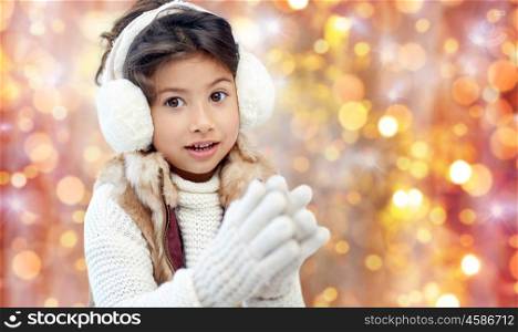 winter, people, christmas and clothing concept - happy little girl wearing earmuffs and gloves over golden holidays lights background