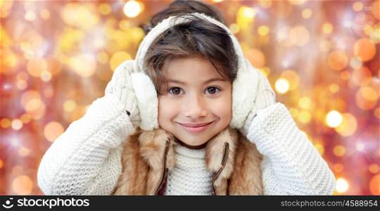 winter, people, christmas and clothing concept - happy little girl wearing earmuffs and gloves over golden holidays lights background