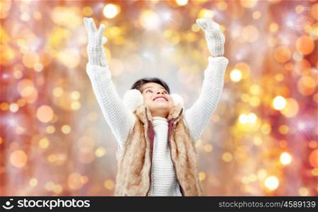 winter, people, christmas and happiness concept - happy little girl wearing earmuffs and gloves over holidays lights background