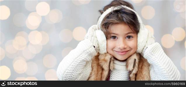 winter, people, christmas happiness concept - happy little girl wearing earmuffs and gloves over holidays lights background