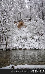 Winter snowy day near a small river. Snow covered forest, dark river water.