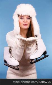 Winter sport activity concept. Atractive girl making kiss gesture wearing warm clothes, ice skates and furry hat, blue background studio shot.. Woman with ice skates kiss gesture