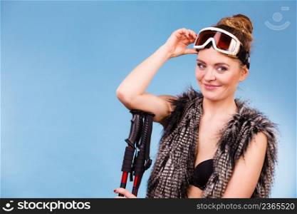Winter sport activity concept. Atractive girl wearing black bra, ski goggles and furry waistcoat holding ski poles under elbow, blue background studio shot.. Woman holding ski goggles and poles