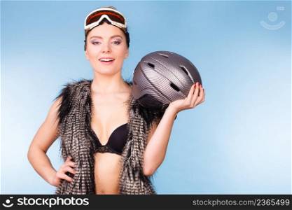 Winter sport activity concept. Atractive smiling girl wearing black bra, ski goggles and furry waistcoat holding helmet, blue background studio shot.. Woman wearing bra and holding ski helmet
