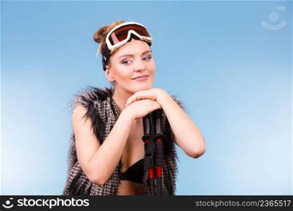 Winter sport activity concept. Atractive smiling girl wearing black bra, ski goggles and furry waistcoat holding ski poles, blue background studio shot.. Sexy woman holding ski poles