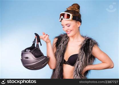 Winter sport activity concept. Atractive smiling girl wearing black bra, ski goggles and furry waistcoat holding helmet, blue background studio shot.. Woman wearing bra and holding ski helmet