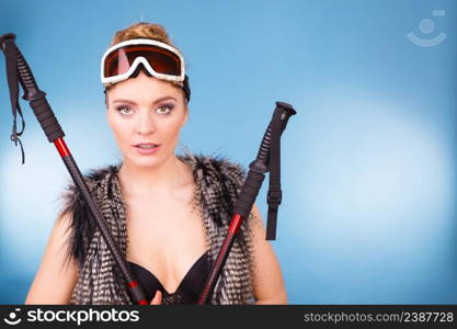 Winter sport activity concept. Atractive smiling girl wearing black bra, ski goggles and furry waistcoat holding ski poles, blue background studio shot.. Sexy woman holding ski poles