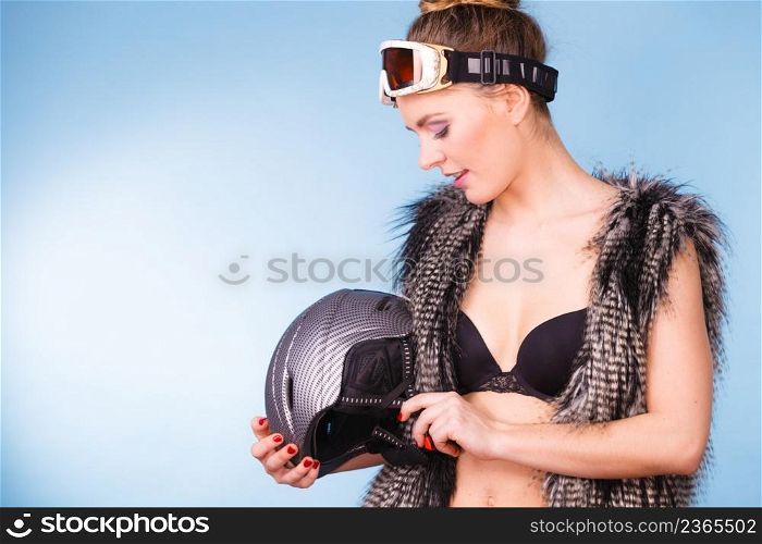Winter sport activity concept. Atractive smiling woman wearing black bra, ski goggles and furry waistcoat holding helmet, blue background studio shot.. Woman wearing sexy winter sport outfit