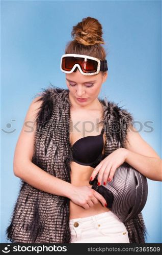 Winter sport activity concept. Atractive smiling woman wearing black bra, ski goggles and furry waistcoat holding helmet, blue background studio shot.. Woman wearing sexy winter sport outfit