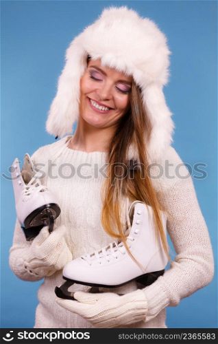 Winter sport activity concept. Girl wearing warm hat and cozy white clothes holding and looking at ice skate, blue background studio shot.. Woman wearing winter hat looking at ice skate