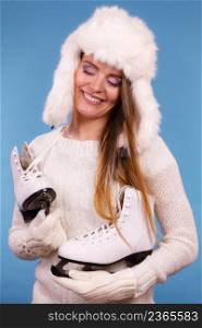 Winter sport activity concept. Girl wearing warm hat and cozy white clothes holding and looking at ice skate, blue background studio shot.. Woman wearing winter hat looking at ice skate