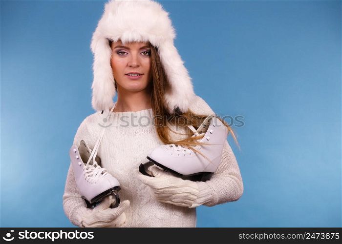 Winter sport activity concept. Girl wearing warm hat and cozy white clothes holding ice skate, blue background studio shot.. Woman wearing winter hat holding ice skate