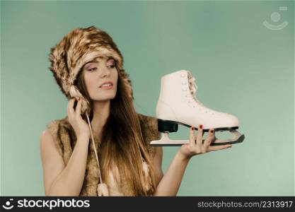 Winter sport activity concept. Girl wearing warm hat and furry waistcoat holding and looking at ice skate, green background studio shot.. Woman wearing winter hat looking at ice skate