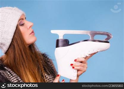 Winter sport activity concept. Girl wearing warm hat and furry waistcoat holding and looking at ice skate, blue background studio shot.. Woman wearing winter hat looking at ice skate