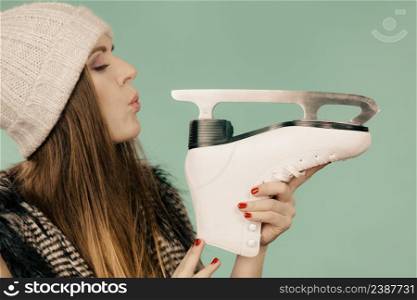 Winter sport activity concept. Girl wearing warm hat and furry waistcoat holding and looking at ice skate, green background studio shot.. Woman wearing winter hat looking at ice skate