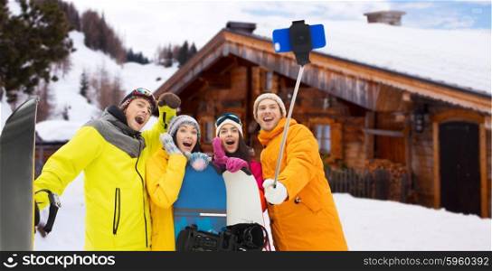 winter sport, leisure, friendship, technology and people concept - happy friends with snowboards and taking picture by smartphone on selfie stick over wooden country house background and snow