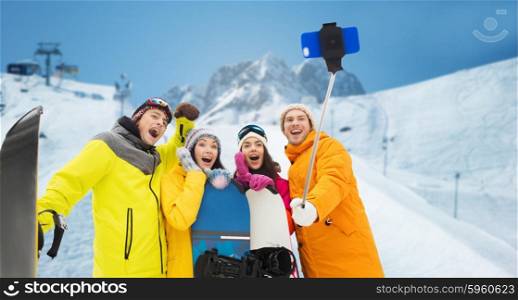 winter sport, leisure, friendship, technology and people concept - happy friends with snowboards and taking picture by smartphone on selfie stick over downhill skiing and mountains background
