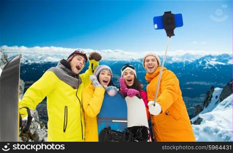 winter sport, leisure, friendship, technology and people concept - happy friends with snowboards and taking picture by smartphone on selfie stick over snowy mountains background