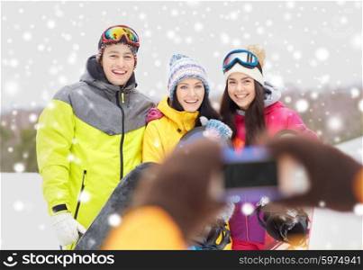 winter sport, leisure, friendship, technology and people concept - happy friends with snowboards and smartphone taking picture