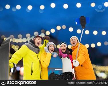 winter sport, technology and people concept - happy friends with snowboards and taking picture by smartphone on selfie stick over christmas lights background. happy friends with snowboards and smartphone
