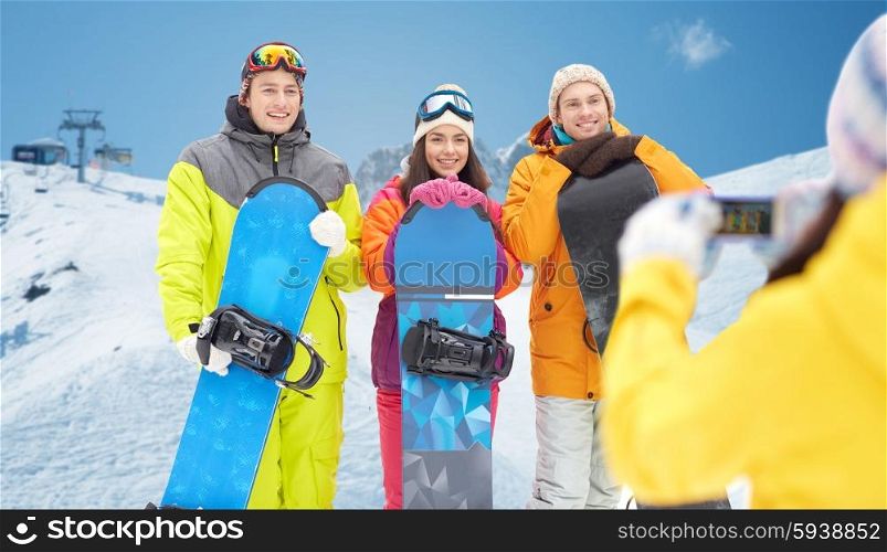 winter sport, technology, leisure, friendship and people concept - happy friends with snowboards and smartphone taking picture over snow and mountain background