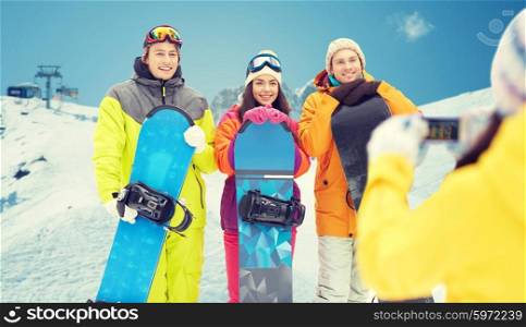 winter sport, technology, leisure, friendship and people concept - happy friends with snowboards and smartphone taking picture over snow and mountain background