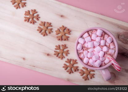 Winter sweets in pink shades, a cup of hot chocolate with mini marshmallows and a Christmas candy cane and snowflakes shaped gingerbread cookies.