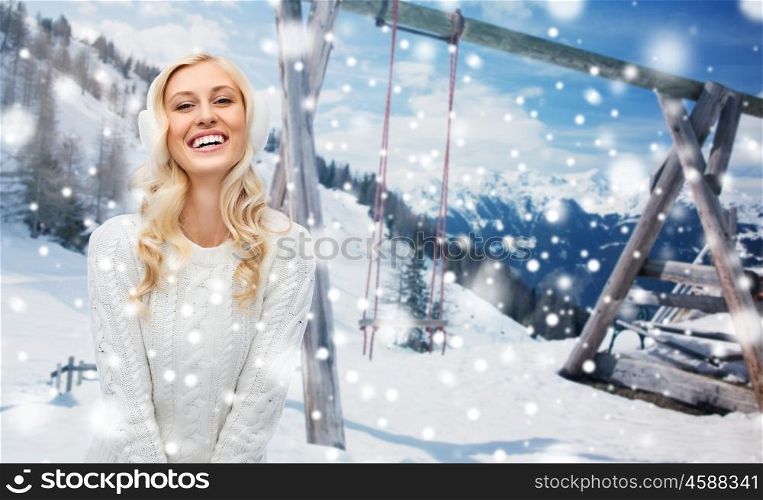 winter, vacation, christmas and people concept - smiling young woman in earmuffs and sweater over snowy mountains and wooden swing background